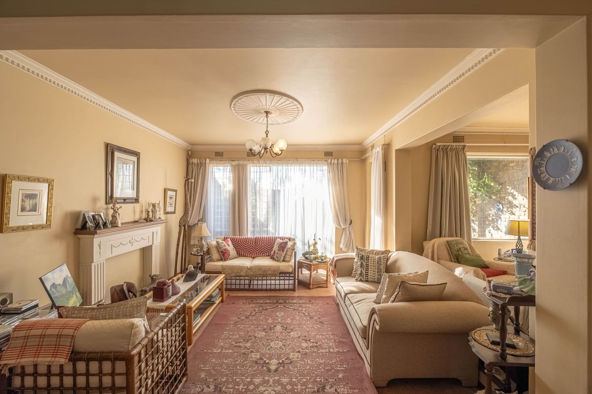 A traditional living room featuring a fireplace mantel, decorative ceiling rose, and crown molding, with large windows providing natural light.