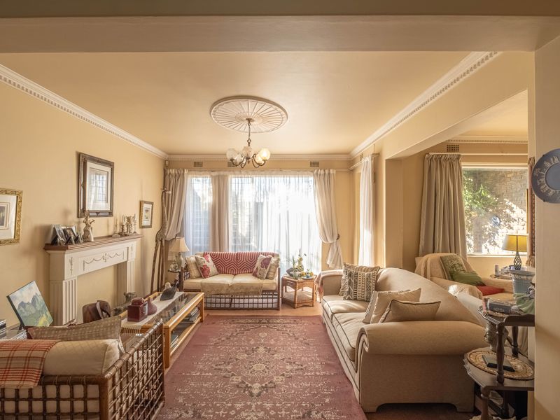 A traditional living room featuring a fireplace mantel, decorative ceiling rose, and crown molding, with large windows providing natural light.