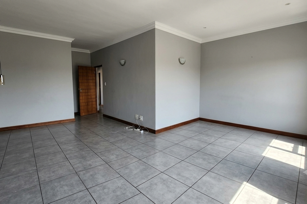 A spacious, empty living area featuring neutral grey walls, tiled flooring, coving, and modern wall-mounted lighting fixtures.