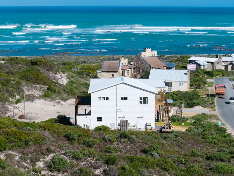 Aerial view of a coastal property featuring a white exterior, metal roofing, and timber decking, situated near the ocean.