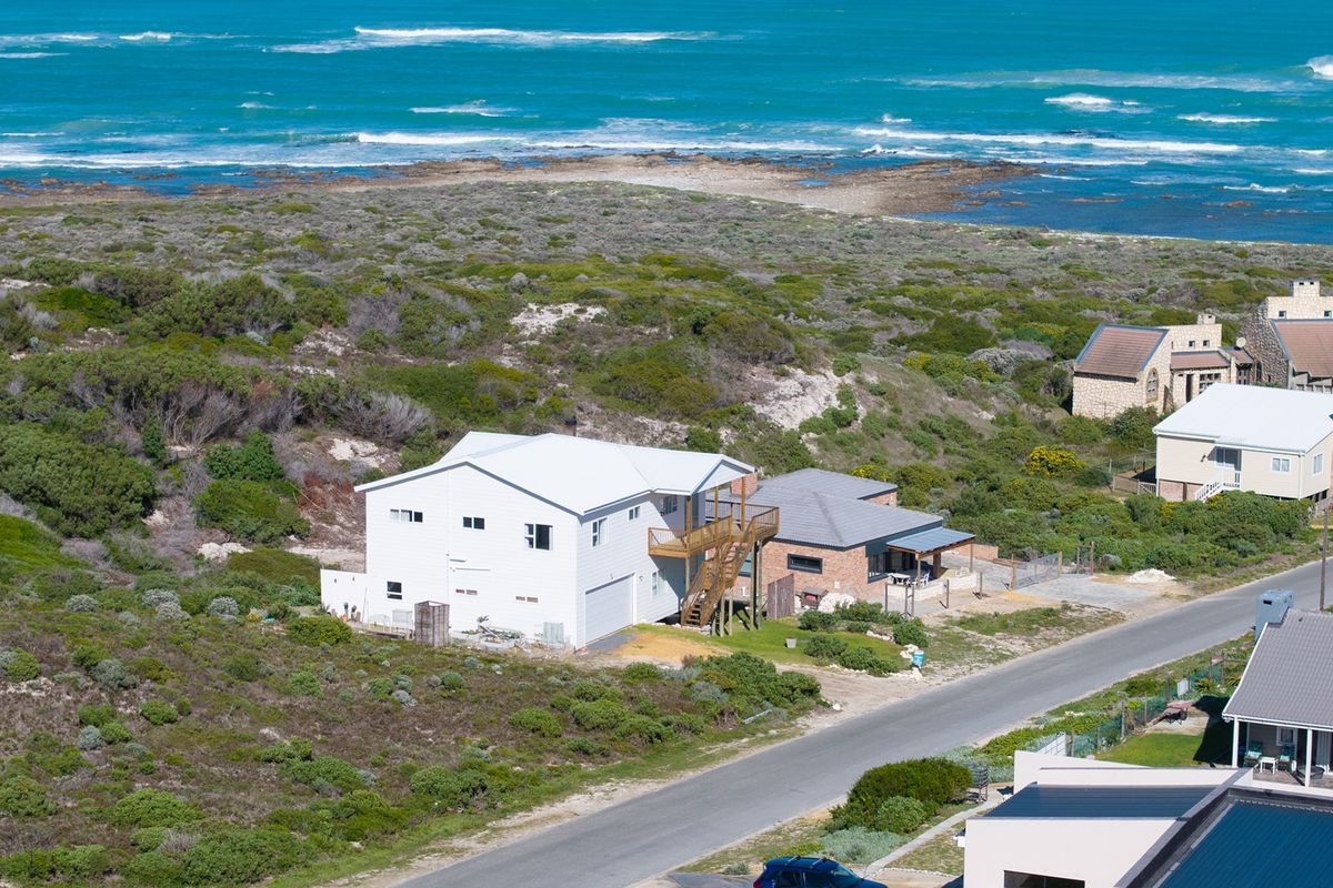 Aerial view of a coastal property featuring a white multi-level home with a metal roof, timber deck, and external staircase, situated near the ocean.