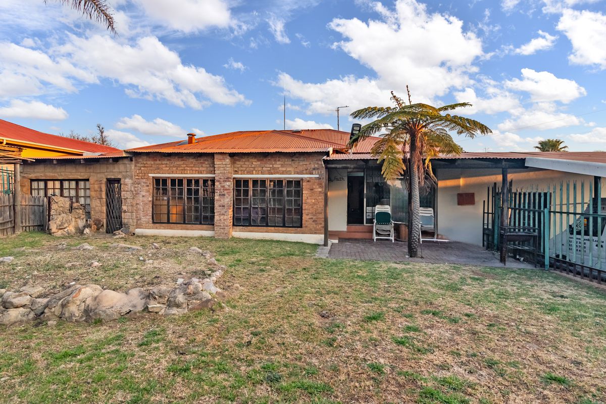Rear view of a brick house featuring a corrugated metal roof, timber-framed windows, and a paved patio area overlooking a garden.