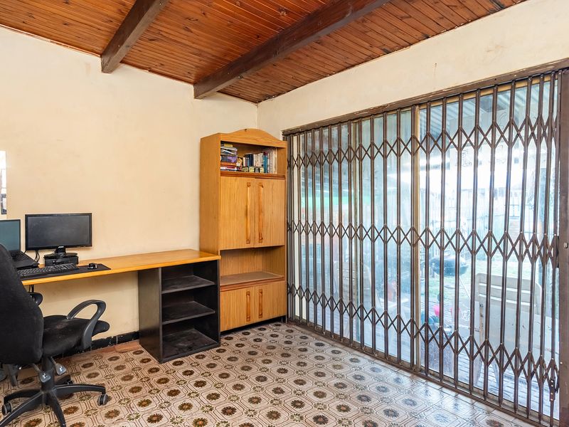 A home office space featuring exposed timber ceiling beams, patterned ceramic flooring, and a large sliding glass door with a protective metal security gate.