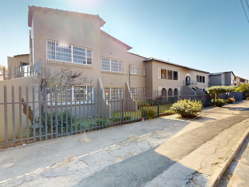 Street-level view of the townhouse complex facade, featuring security fencing, security bars on windows, and a tiled roof structure.