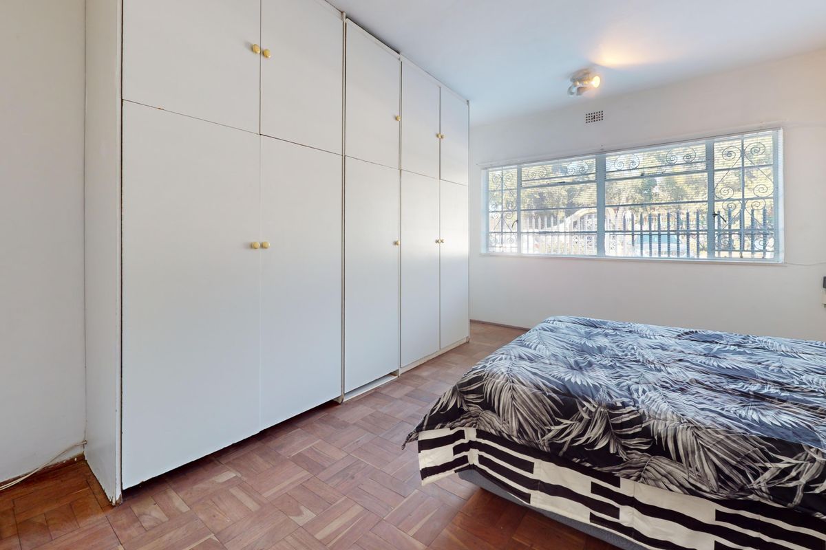 A bedroom featuring parquet flooring, extensive built-in cupboards for storage, and a window fitted with security bars.
