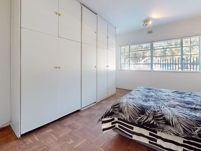 A bedroom featuring parquet flooring, extensive built-in cupboards for storage, and a window fitted with security bars.