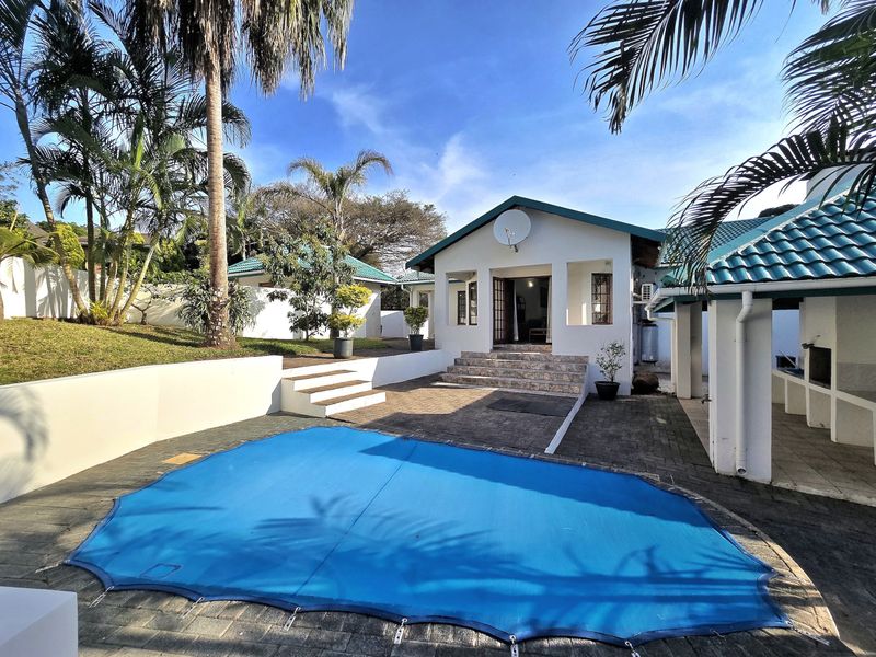 Outdoor pool area featuring a paved patio, built-in braai, and a covered swimming pool, set against a well-maintained garden and residential building.