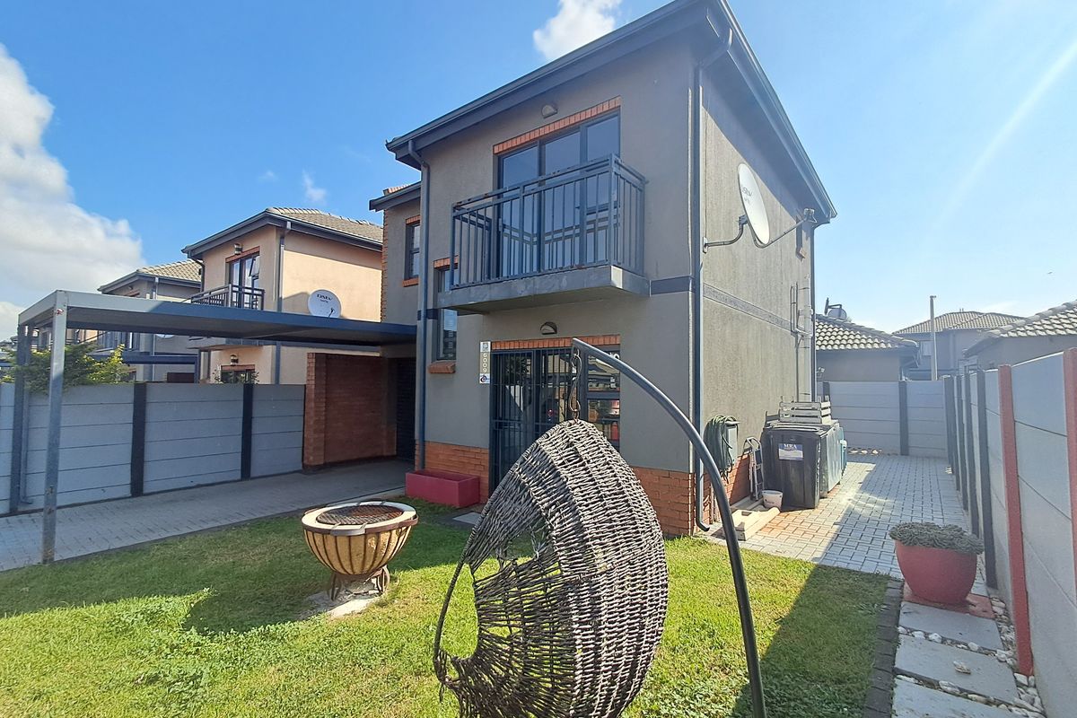 Rear view of a two-story residential property featuring a paved driveway, carport, small garden area, and an upper-level balcony with metal railings.