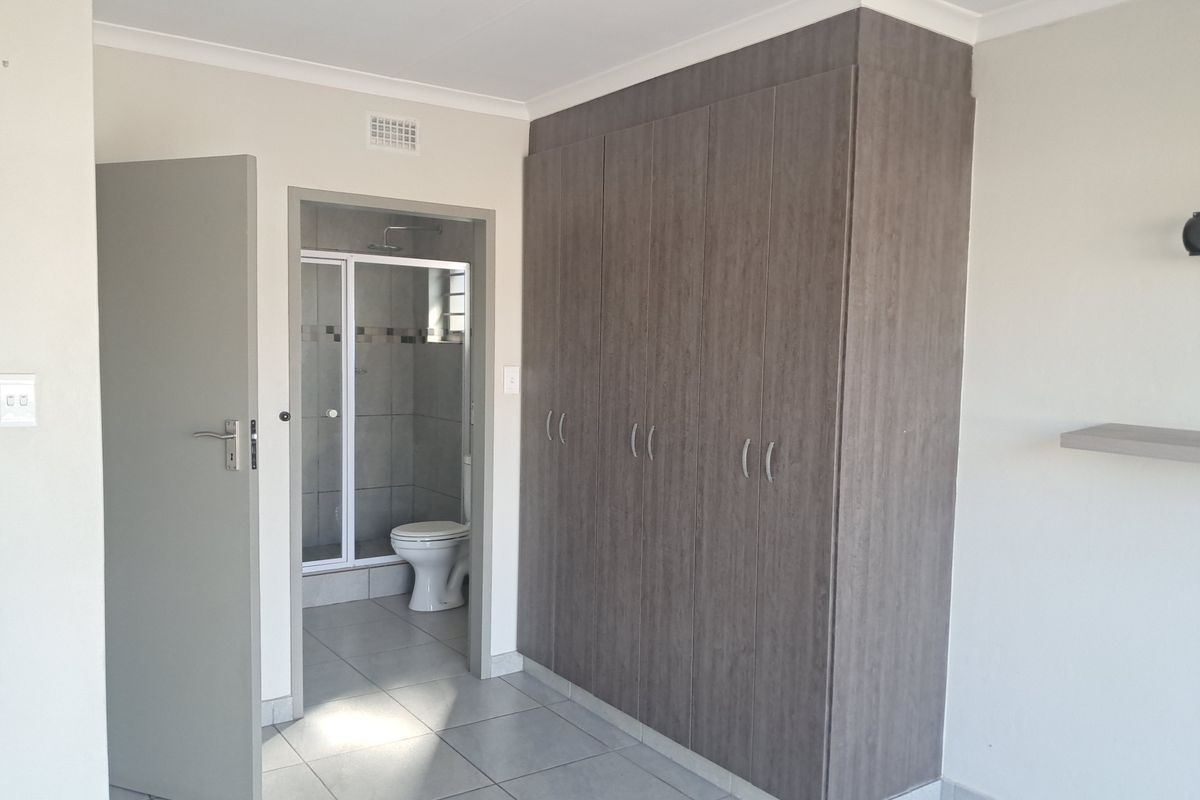A clean, modern bedroom featuring neutral tiled flooring, ample built-in cupboard storage, and an adjoining en-suite bathroom with a shower.