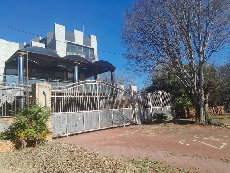 Front view of a multi-story residence featuring a large metal security gate, paved driveway, and an elevated covered patio area with glass balustrades.