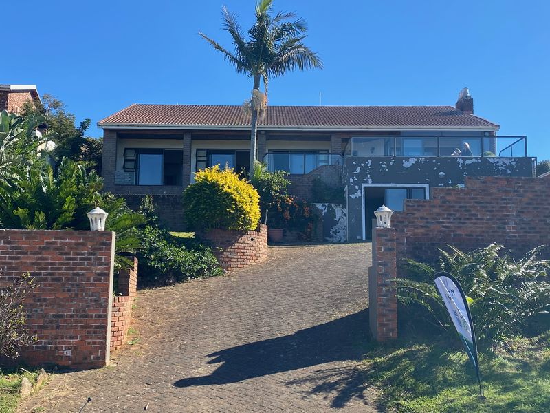 Front elevation of a multi-level brick house featuring a paved driveway, balcony with glass balustrade, and mature garden landscaping.