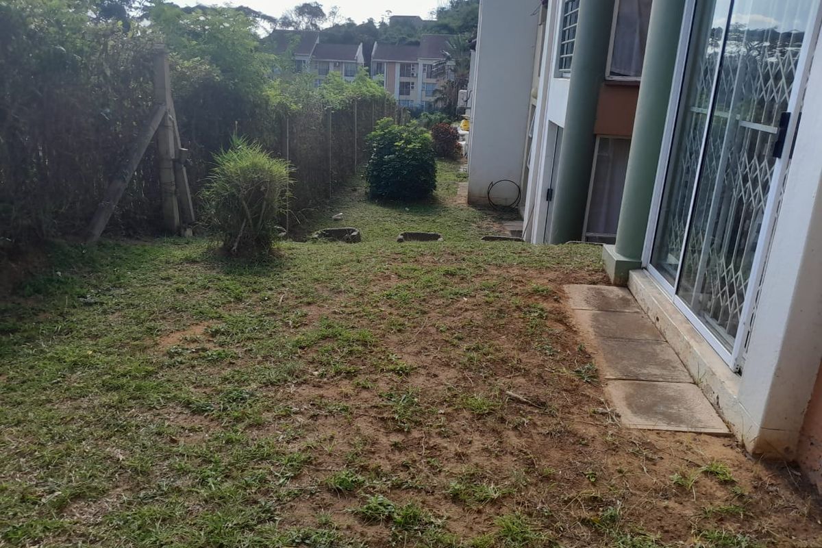 A rear garden area featuring a patch of lawn, perimeter fencing, and access to the apartment via sliding glass doors with security gates.