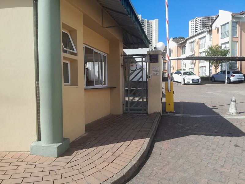 Security entrance and guard booth for the apartment complex, featuring a boom gate, pedestrian access, and paved driveway area.