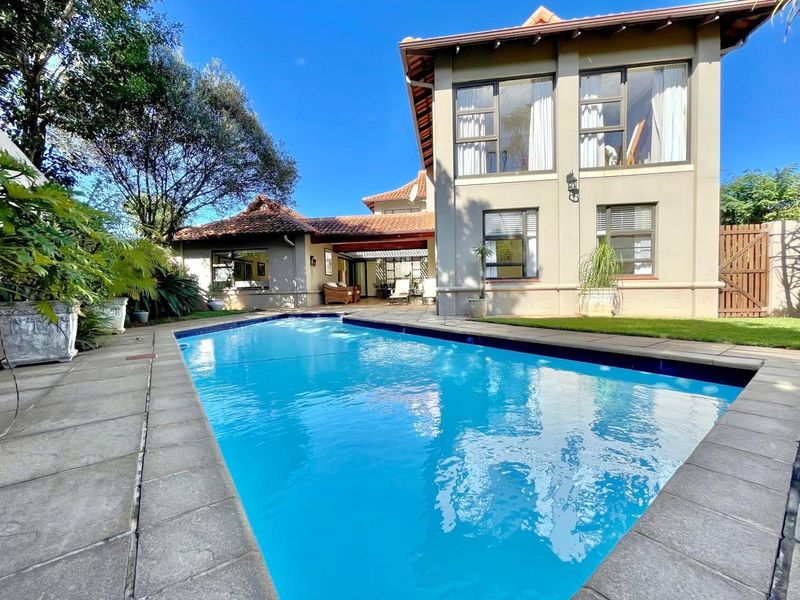 A well-maintained swimming pool area featuring a paved deck, covered patio, and a two-story residential structure with large windows.
