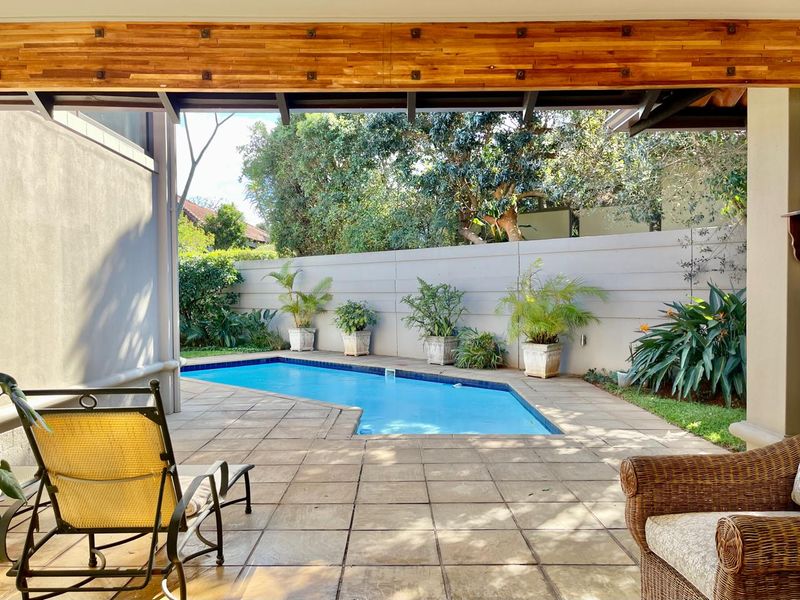 A private outdoor pool area featuring a tiled patio, swimming pool, and a covered timber-beamed patio space enclosed by a boundary wall.
