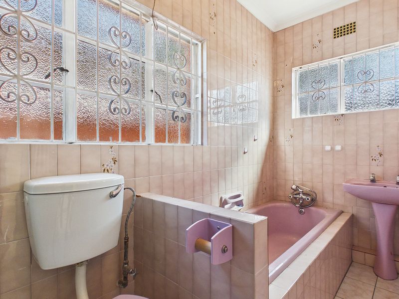 A dated bathroom featuring original pink sanitaryware, tiled walls, and security bars on the windows, offering significant potential for a modern renovation.
