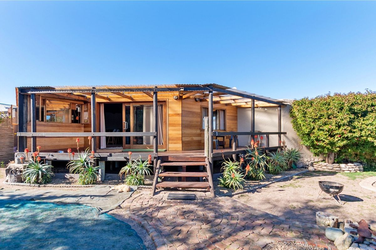 Rear view of a timber-clad dwelling featuring a raised wooden deck, pergola, and sliding door access, overlooking a garden area with paved pathways.