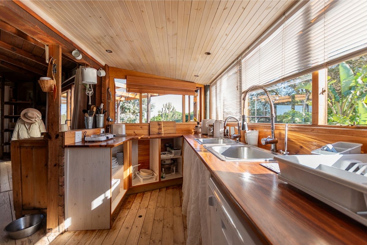 A rustic kitchen featuring timber wall cladding, wooden countertops, a double stainless steel sink with a gooseneck faucet, and large windows with venetian blinds.
