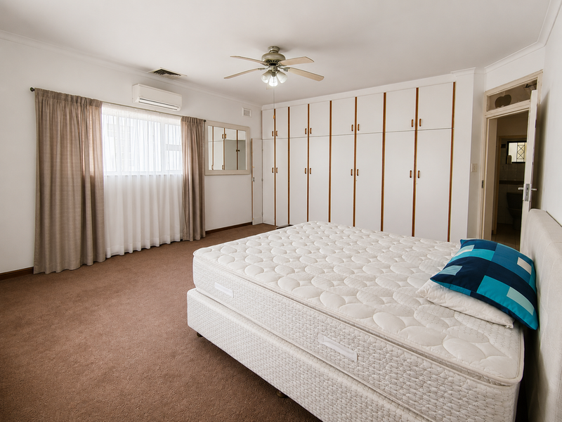 Spacious bedroom featuring extensive built-in cabinetry, carpeted flooring, a ceiling fan, and a wall-mounted air conditioning unit with an adjoining en-suite bathroom.