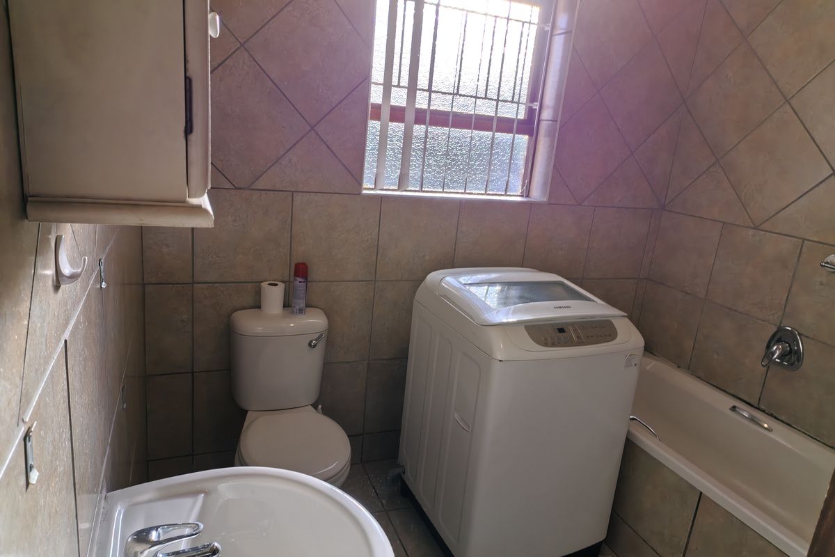 A functional bathroom featuring a bathtub, pedestal basin, and toilet, finished with full-height wall tiling and a window with security bars.