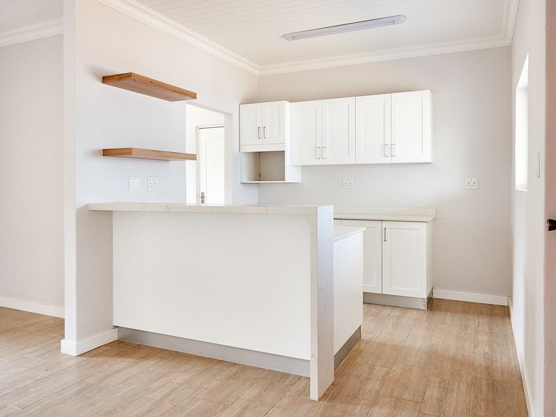 A bright, modern kitchen featuring white cabinetry, stone countertops, a breakfast bar, and timber floating shelves, finished with neutral tiled flooring.