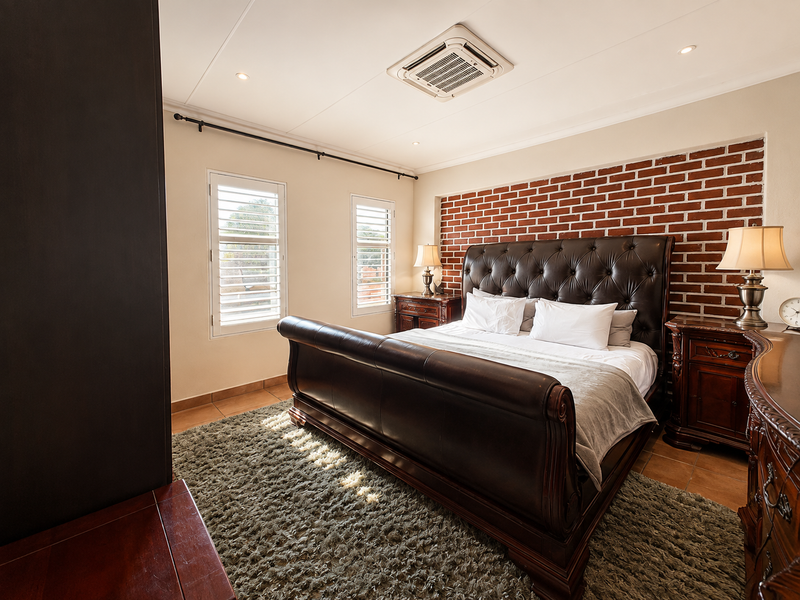 Spacious bedroom featuring an exposed brick accent wall, ceiling-mounted air conditioning, plantation shutters, and tiled flooring.
