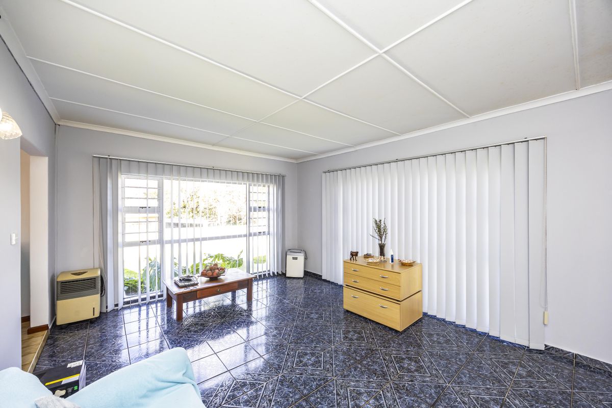 A spacious living area featuring dark patterned floor tiles, large windows with vertical blinds, and a suspended ceiling.