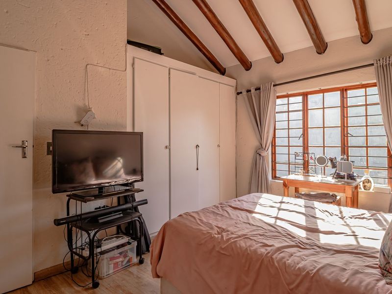 A cozy bedroom featuring exposed timber ceiling beams, built-in cupboards, and a window with security bars, offering a warm and rustic aesthetic.