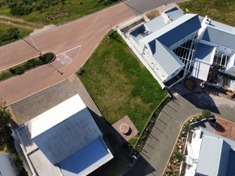An aerial view of a vacant residential plot situated between existing properties, featuring a grass lawn and adjacent paved street access.
