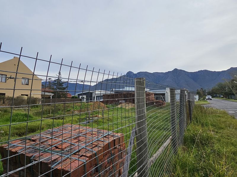 A street-level view of a vacant residential plot featuring wire mesh fencing, adjacent to a paved road with scenic mountain views in the background.