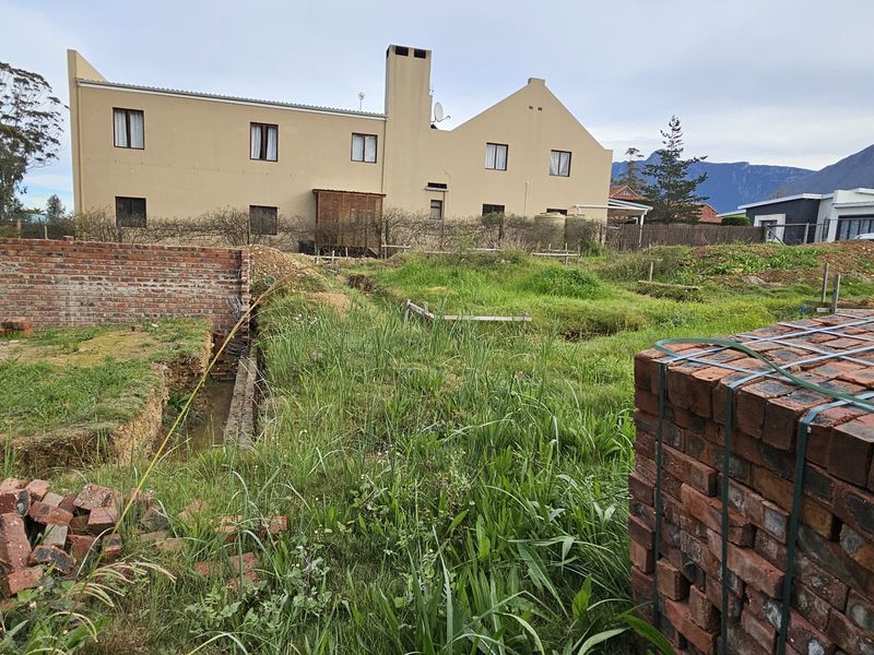 A vacant residential plot featuring ongoing foundation work, brick retaining walls, and stacked construction materials, set against a backdrop of neighboring properties.