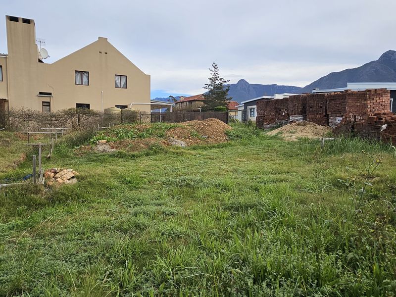 A vacant residential plot featuring overgrown grass, piles of construction bricks, and surrounding neighborhood views.