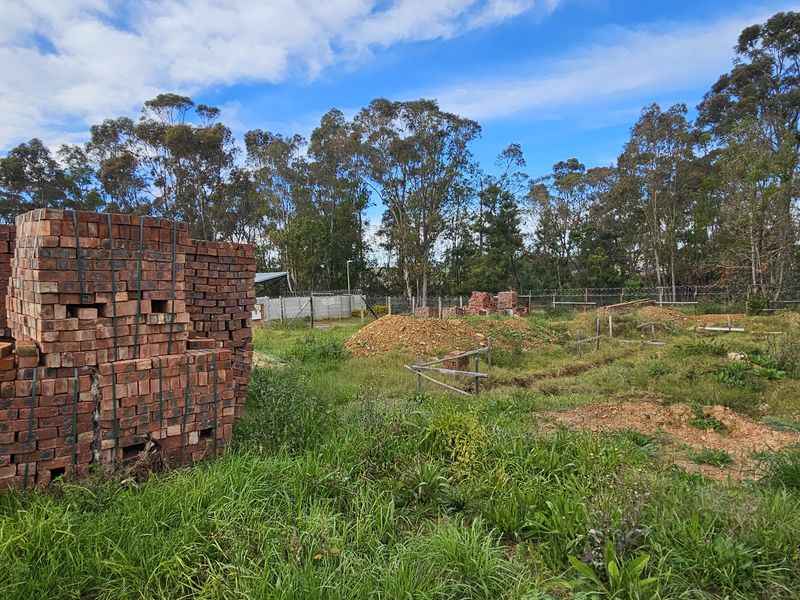 A vacant residential plot featuring cleared land, excavated trenches, and stacked construction bricks, enclosed by wire mesh fencing and wooden posts.