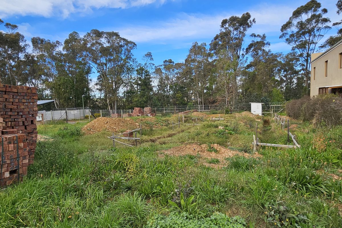 A vacant residential plot featuring excavated foundation trenches, perimeter fencing, and stacked construction bricks, surrounded by established trees.