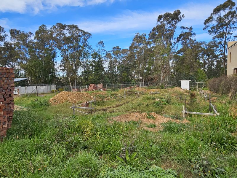 A vacant residential plot featuring excavated foundation trenches, perimeter fencing, and stacked construction bricks, surrounded by established trees.