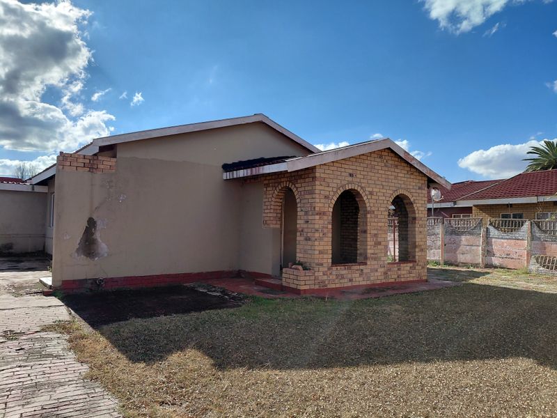 Front elevation of a single-story house featuring an arched brick porch, paved driveway, and boundary wall, showing signs of exterior maintenance requirements.