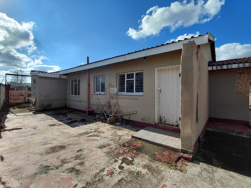 Rear exterior view of the house featuring a concrete patio area, security gate, and exposed plumbing, showing significant signs of wear and maintenance requirements.