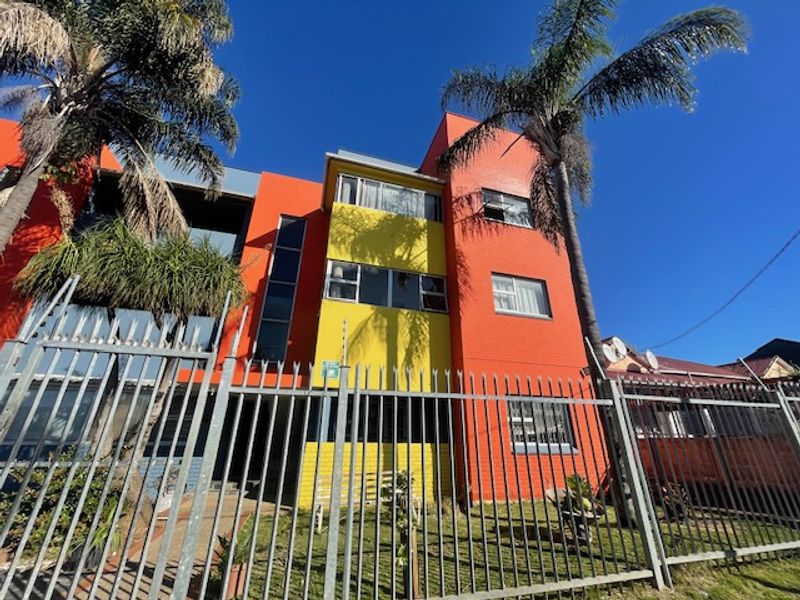 Front elevation of a multi-story apartment building featuring a vibrant orange and yellow painted facade with secure metal perimeter fencing.