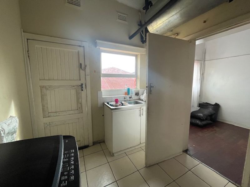 A functional scullery area featuring a stainless steel sink, tiled flooring, and exposed overhead plumbing, offering potential for modernization and improved utility.