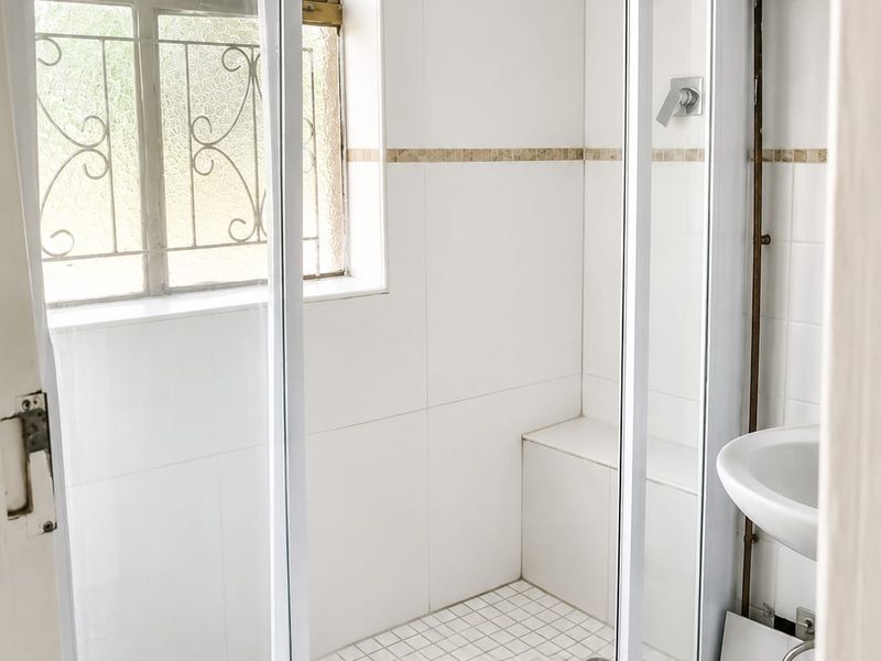 A functional bathroom featuring a walk-in shower with a built-in bench, white wall tiles, and a pedestal basin with exposed plumbing.