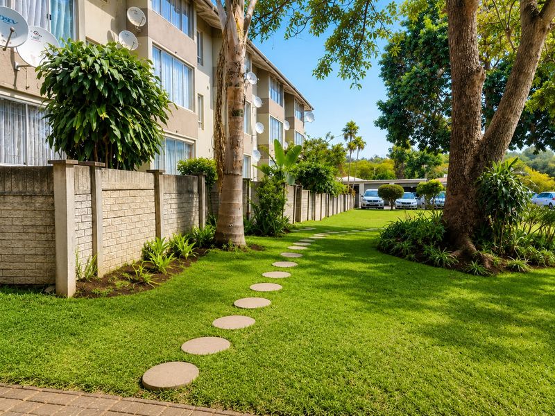 A well-maintained communal garden area featuring a stone pathway, mature trees, and a concrete block boundary wall alongside the apartment building.