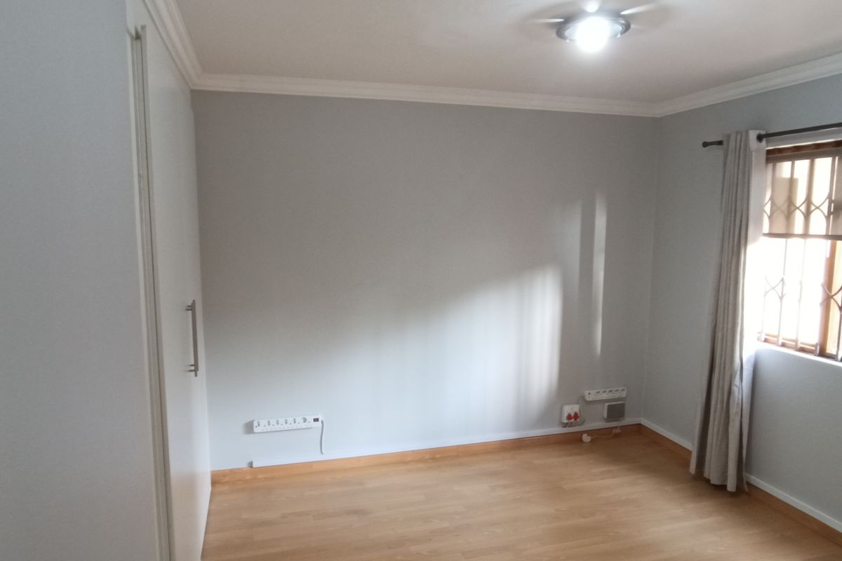 A clean, empty bedroom featuring laminate flooring, built-in cupboards, and a window fitted with a security gate.