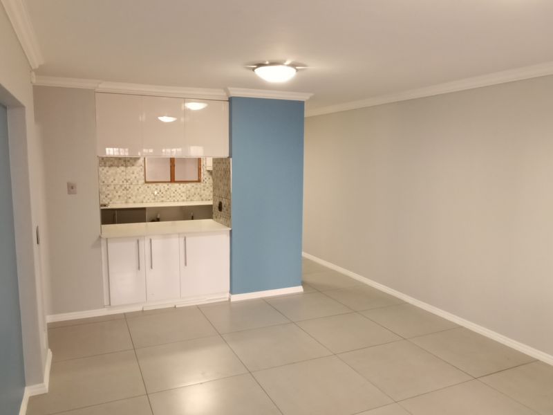 An open-plan living area featuring tiled flooring, neutral walls with a blue accent, and a compact kitchen corner with built-in cabinetry and mosaic backsplash.