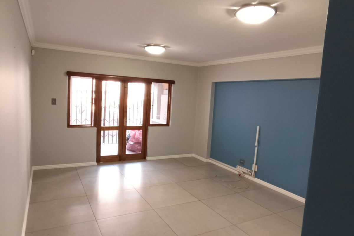 A bright, empty living room featuring large format floor tiles, neutral wall tones, and sliding glass doors with security gates leading to the exterior.