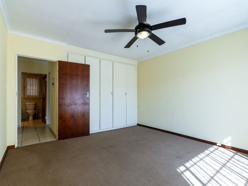 A spacious bedroom featuring built-in cupboards, a ceiling fan, and carpeted flooring, with an adjoining bathroom visible through the doorway.