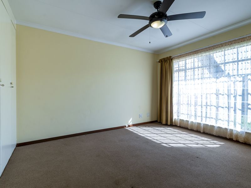 A spacious, empty bedroom featuring neutral walls, carpeted flooring, built-in cupboards, and a ceiling fan, with large windows allowing for ample natural light.