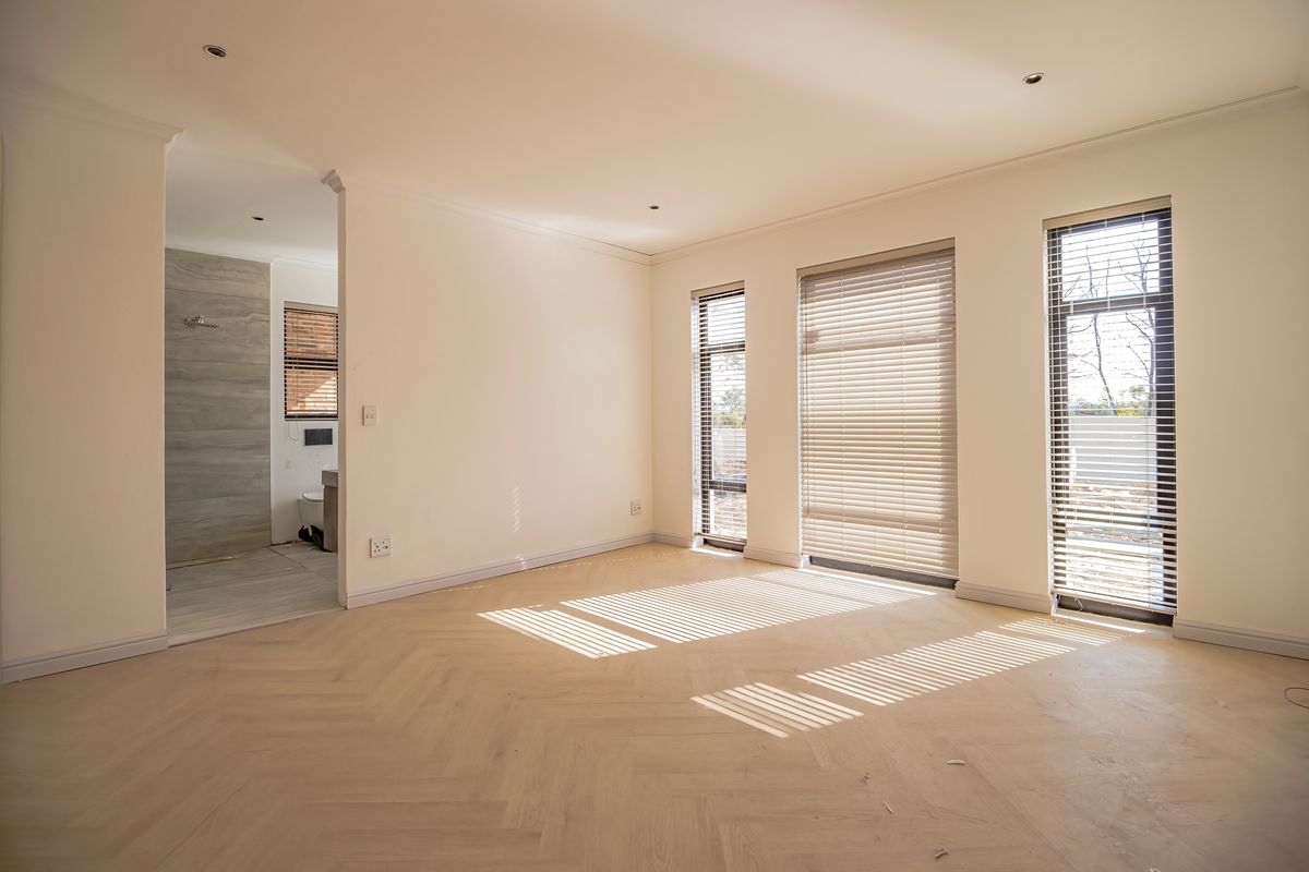 A bright, modern bedroom featuring light herringbone timber flooring, recessed downlights, and large windows with fitted venetian blinds, leading into an en-suite bathroom.