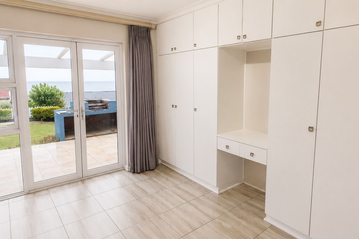 A bright, empty bedroom featuring extensive built-in cabinetry, a vanity area, tiled floors, and direct access to an outdoor patio with garden views.