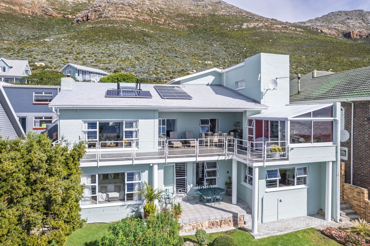 Rear view of a multi-level house featuring a spacious balcony, ground-floor patio, solar panels, and established garden set against a mountain backdrop.