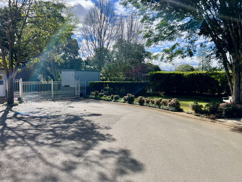 A view of the property entrance featuring an asphalt driveway, metal security gate, and well-maintained landscaped garden borders with mature trees.